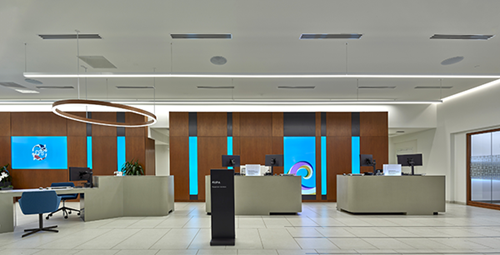 A modern Central Pacific Bank branch interior featuring multiple teller stations, wood-paneled walls, blue accent lighting, and a circular overhead light fixture. The bright, open banking space has a clean contemporary design with service counters arranged across the lobby.