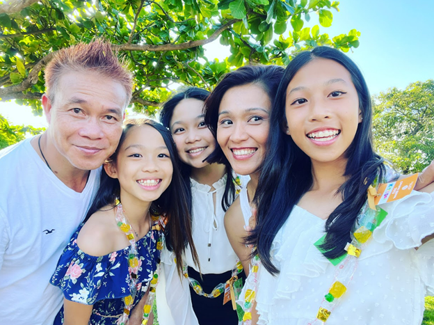 A family of five poses for a close-up outdoor photo beneath a leafy tree canopy. The group stands together wearing colorful leis, with bright sunlight filtering through the greenery in the background.
