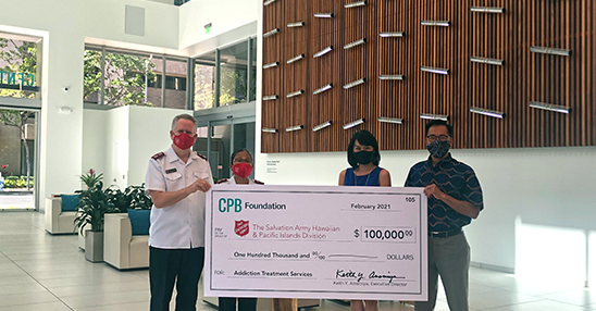 Three people wearing face masks stand in the Central Pacific Bank lobby holding a $100,000 CPB Foundation donation check for The Salvation Army Hawaiian & Pacific Islands Division’s Addiction Treatment Services program. The presentation takes place beneath a decorative wooden wall feature in the bank’s bright, modern lobby.