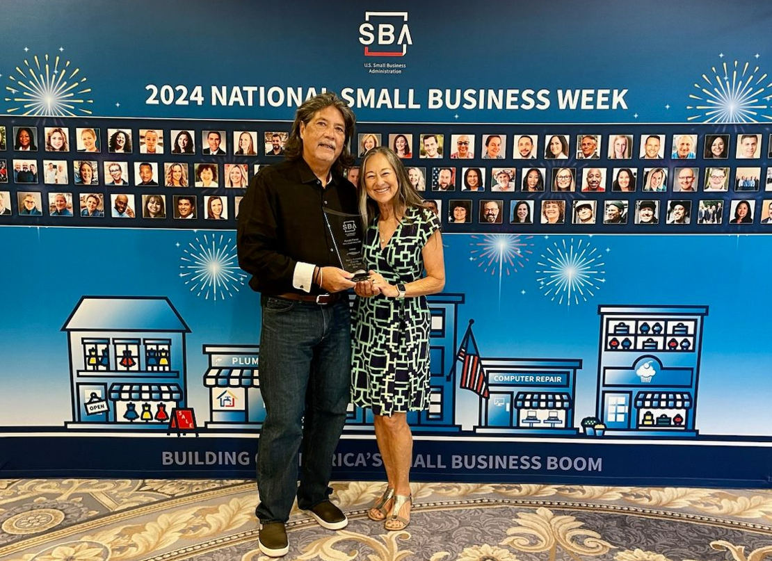 A person receives an award during 2024 National Small Business Week, standing in front of a U.S. Small Business Administration (SBA) display featuring photos of small business owners and illustrated storefronts. The recipient poses with an award plaque against a blue SBA-branded backdrop.