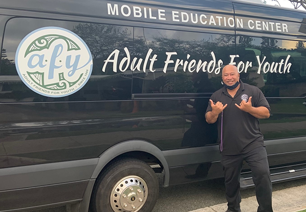 A person stands beside the Adult Friends for Youth Mobile Education Center van, making a shaka gesture. The black vehicle displays the Adult Friends for Youth logo and organization name prominently along the side.