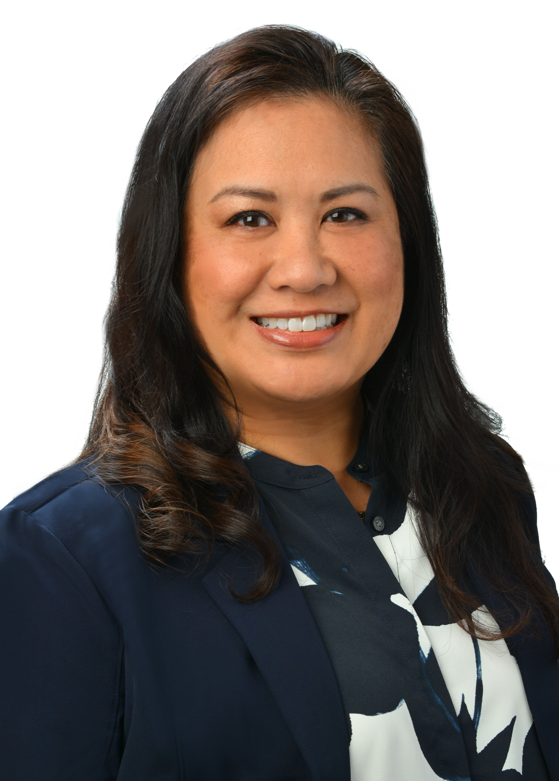 A professional headshot of a person with long dark hair wearing a navy blazer over a patterned blouse, photographed against a light gray background.