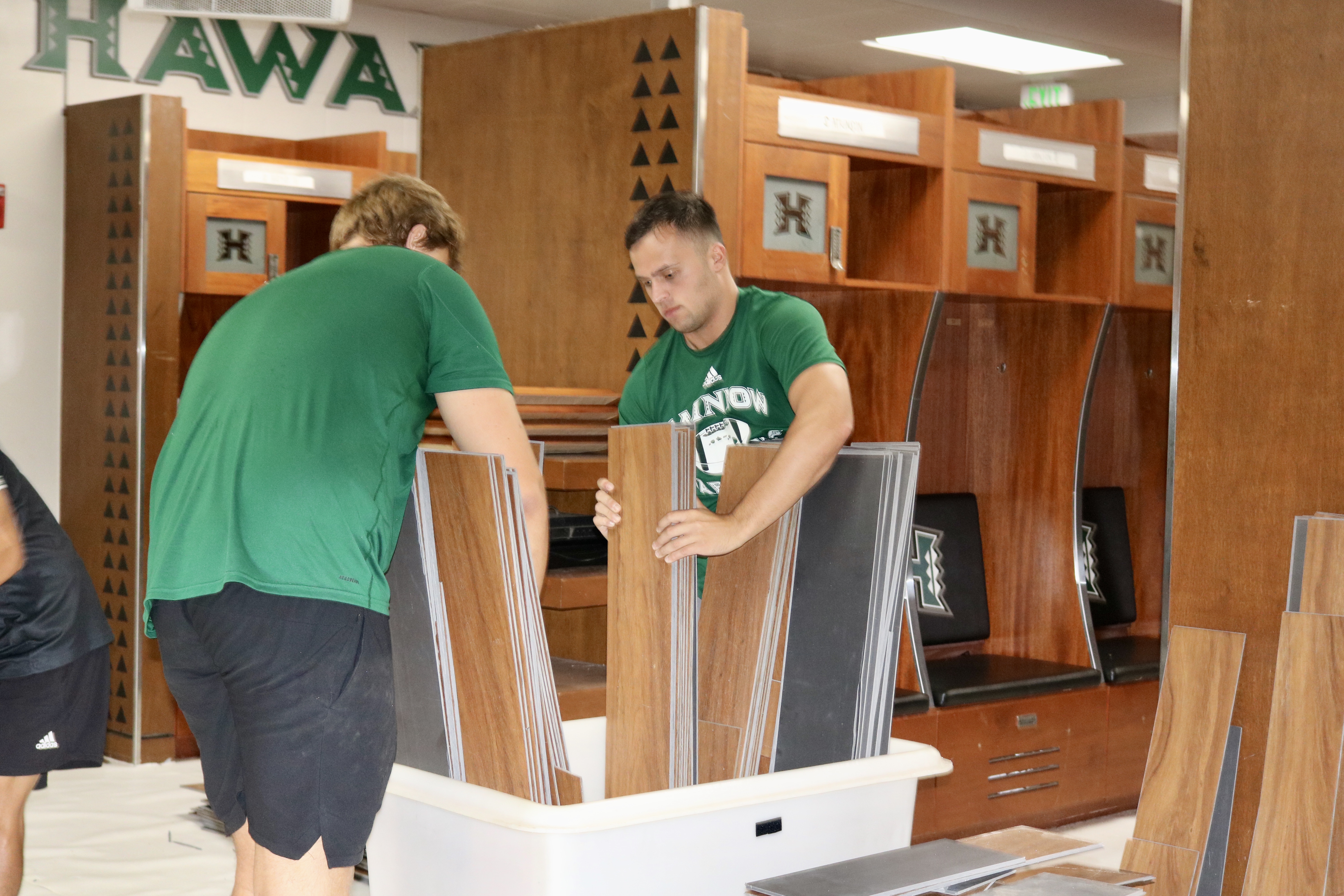 A group of people wearing green University of Hawaiʻi athletics shirts install flooring inside a locker room. Stacks of flooring planks, wooden lockers, and UH-branded seating are visible, highlighting a facility renovation project.
