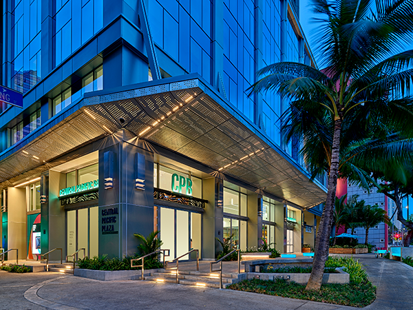 A modern Central Pacific Bank branch is illuminated at dusk, featuring glass storefront windows, contemporary architecture, and prominent CPB signage. Palm trees, landscaped walkways, and the entrance to Central Pacific Plaza frame the building along a downtown city street.