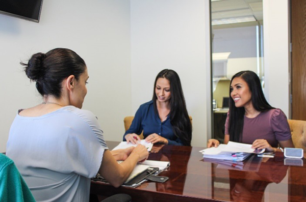 A bank representative meets with two clients at a conference table, reviewing documents during a financial consultation. Papers, a tablet, and office materials are spread across the table in a private meeting room.