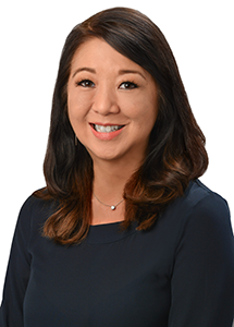 A professional headshot of a person wearing a navy blue blouse and a delicate necklace, photographed against a light gray background.