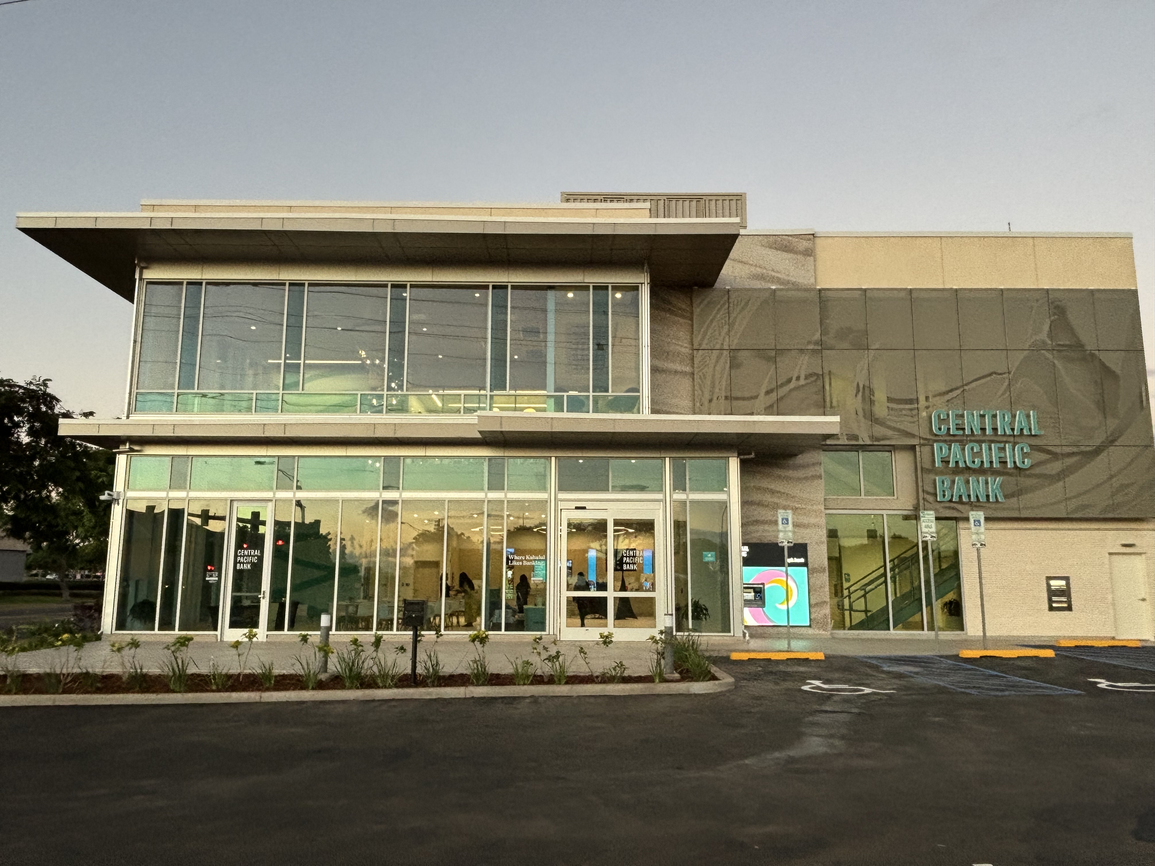 A modern two-story Central Pacific Bank branch features floor-to-ceiling glass windows, prominent teal bank signage, and contemporary architectural details. The building sits behind a newly paved parking area, with illuminated interior spaces visible through the glass façade.