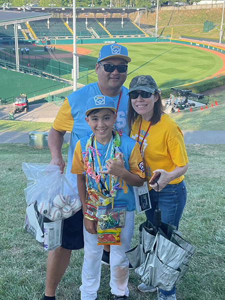 A youth baseball player wearing a team uniform and multiple medals poses with two adults on a grassy hillside overlooking a baseball stadium. The trio stands near the field holding baseballs and event items, with the ballpark and seating visible in the background.