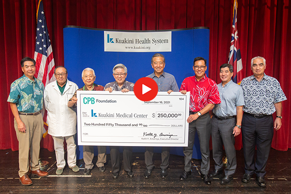A group of seven people stand on a stage holding a large $250,000 donation check for Kuakini Medical Center from the CPB Foundation. The photo is taken in front of a Kuakini Health System backdrop, with U.S. and Hawaii flags positioned on either side of the stage.