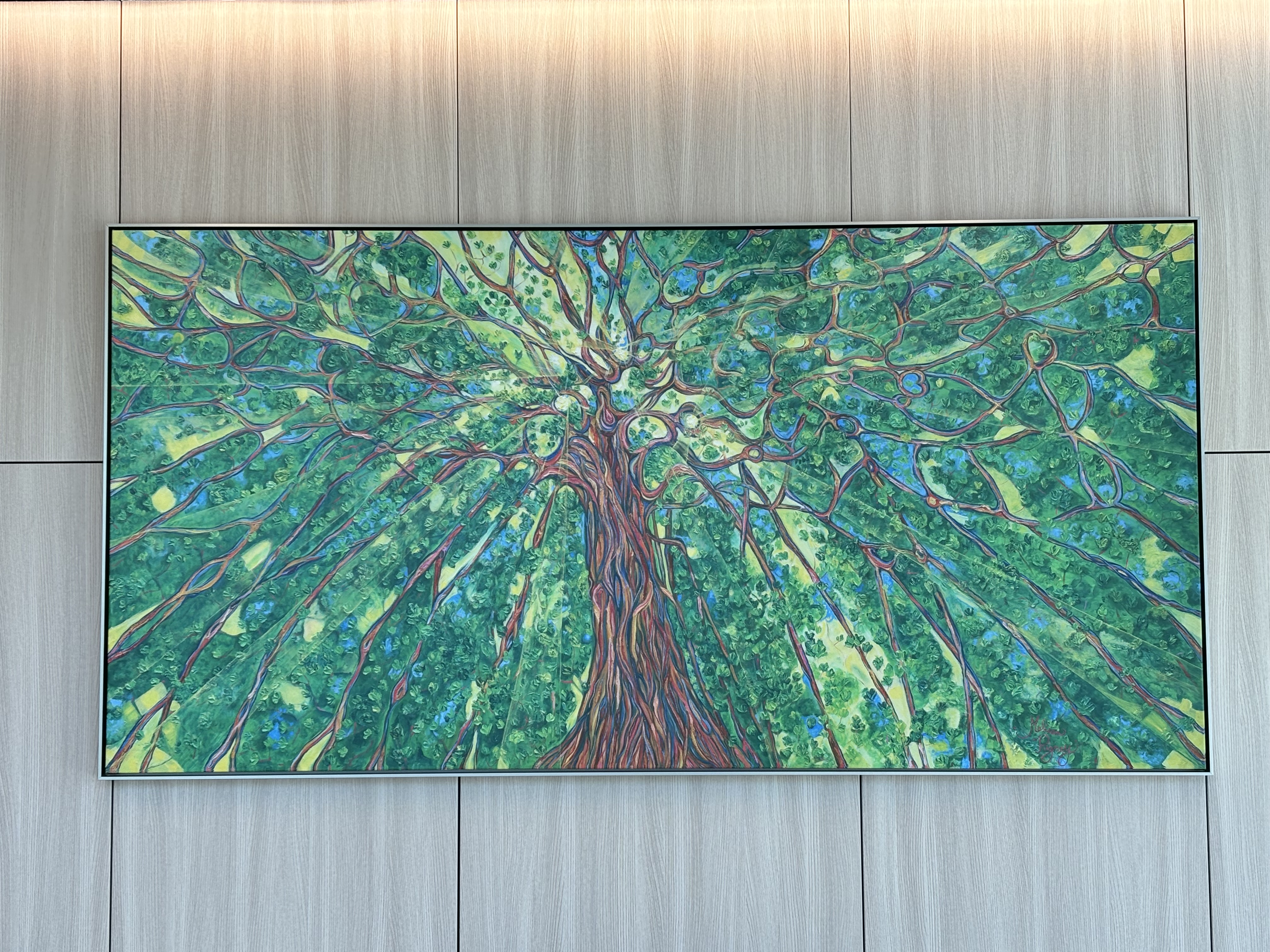 A large colorful painting of a tree viewed from below hangs on a wood-paneled wall. The artwork features a twisting trunk and sprawling green branches that radiate outward across the canvas, creating a vibrant canopy effect.