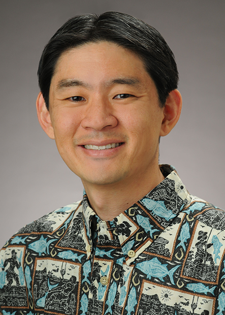 A professional headshot of a person wearing a patterned Hawaiian shirt in shades of blue, black, and beige, photographed against a gray studio background.