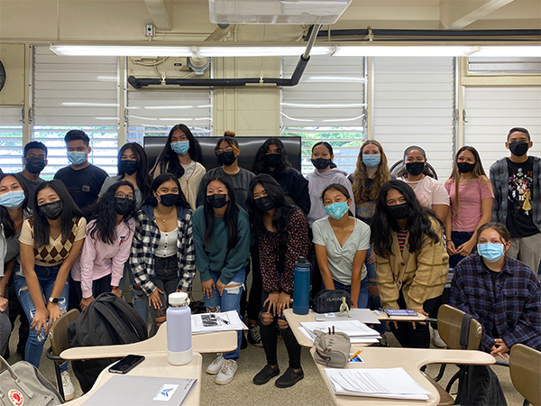 A group of students wearing face masks pose together inside a classroom, standing between rows of desks with notebooks, water bottles, and school supplies visible in the foreground.