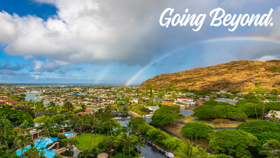 A panoramic view of a Hawaiian community and coastline beneath a rainbow and dramatic clouds, with green palm trees and a sunlit hillside in the foreground. The words “Going Beyond.” appear in the upper-right corner.