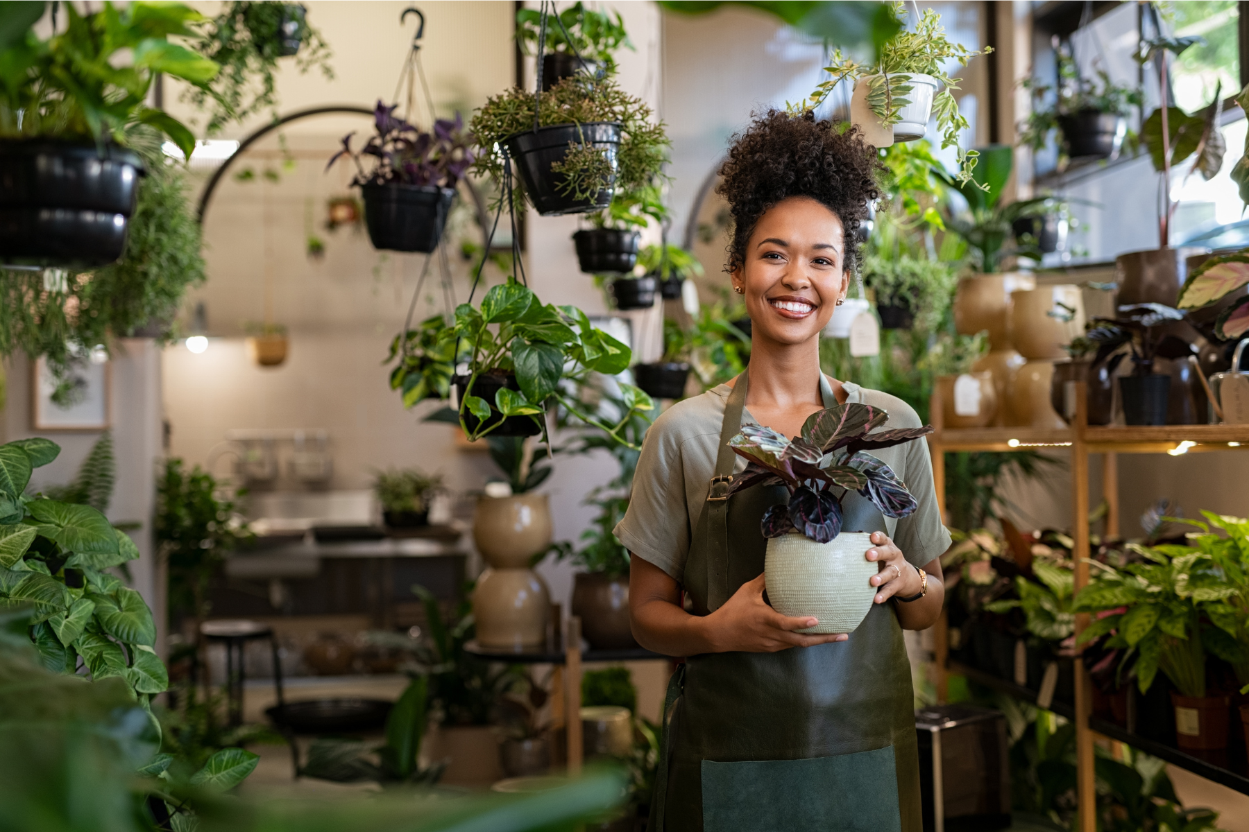 Woman shopkeeper proudly holding a beautiful potted plant (Calathea roseopicta), surrounded by shelves of other well kept plants.