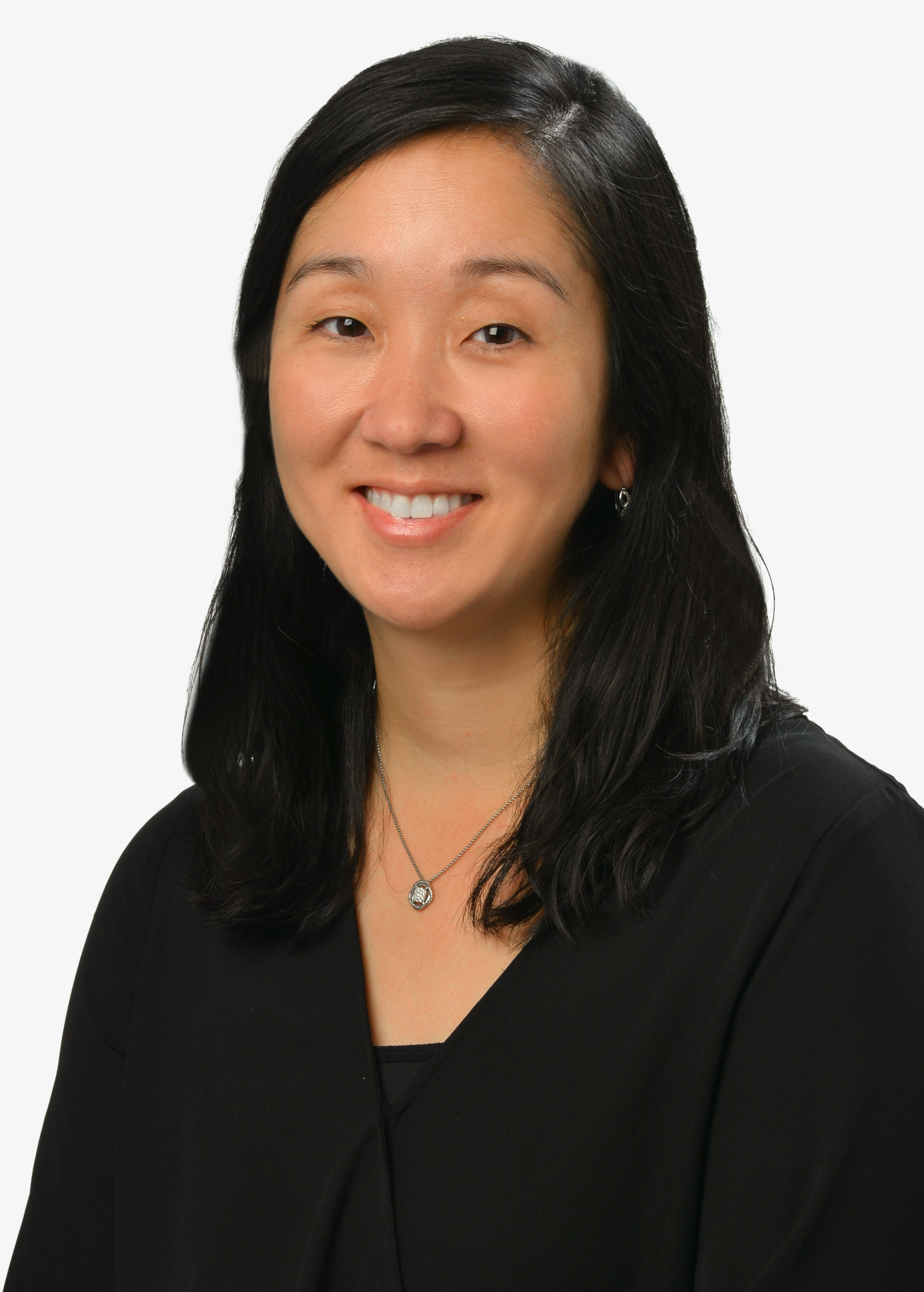 A professional headshot of a person with shoulder-length dark hair wearing a black blouse and a small pendant necklace, photographed against a light gray background.