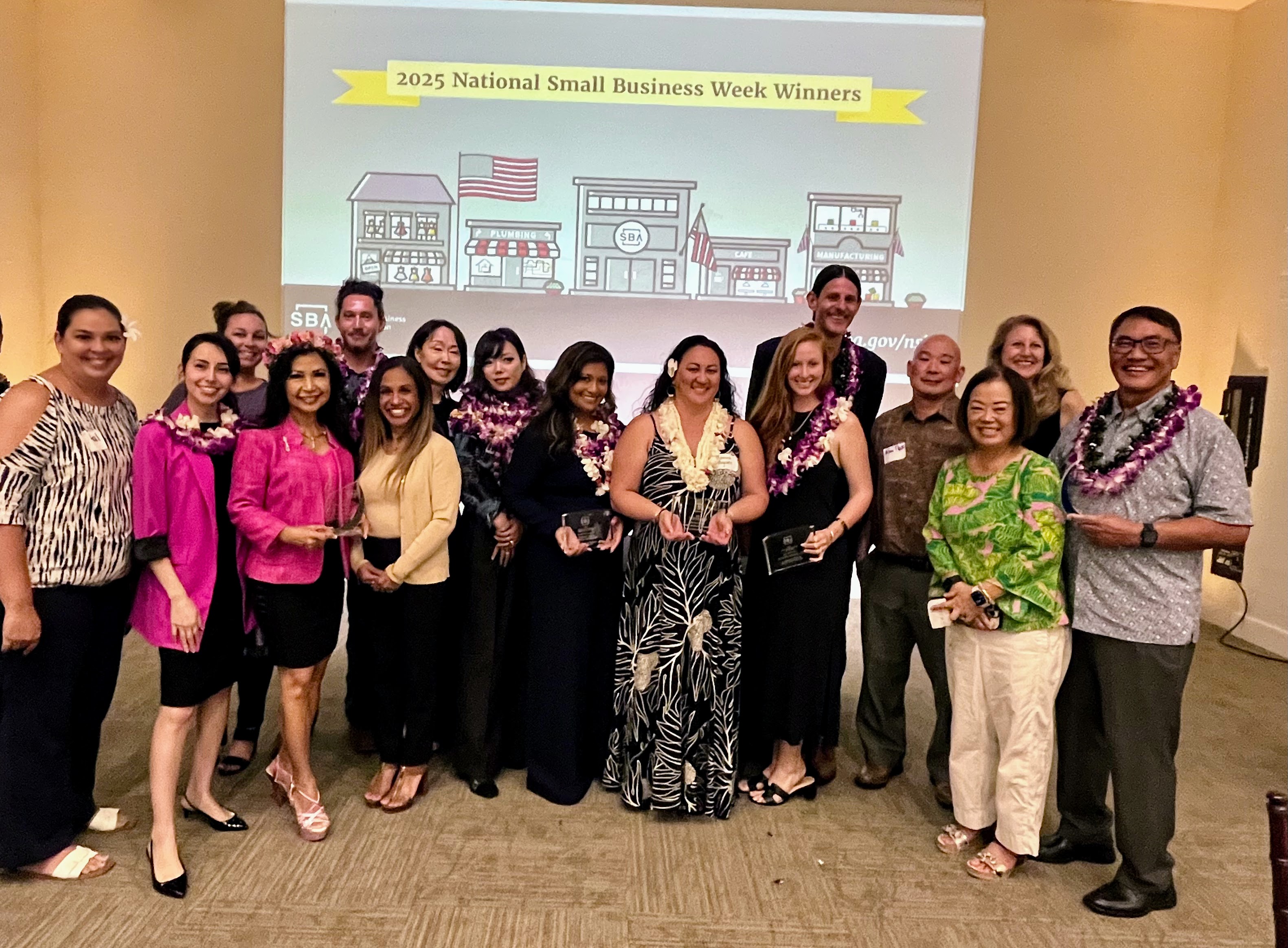 A group of award recipients and community leaders wearing flower leis pose together at a 2025 National Small Business Week Winners recognition event. Several participants hold award plaques while standing in front of a presentation screen displaying SBA branding and National Small Business Week graphics.