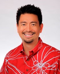 A professional headshot of a person wearing a red aloha shirt with a large white floral pattern, photographed against a light gray background.
