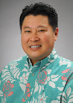 A professional headshot of a person wearing a light blue floral Hawaiian shirt, photographed against a gray studio background.