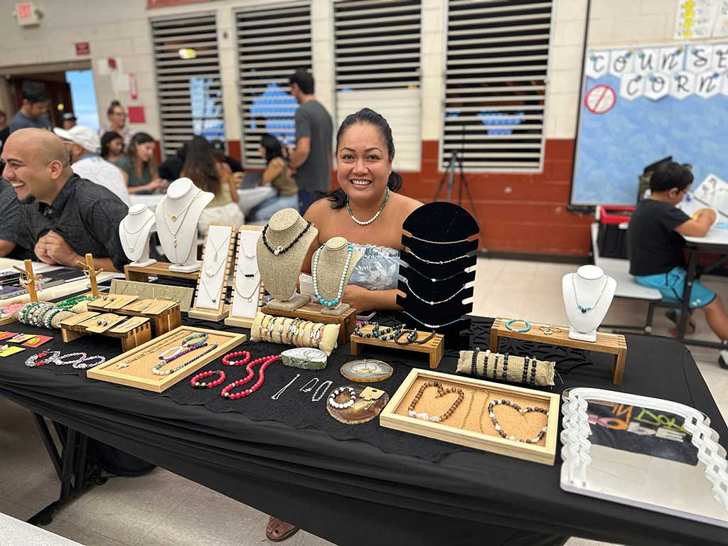 A person sits behind a display table showcasing handmade jewelry, including necklaces, bracelets, and earrings arranged on stands and trays. The booth is set up at an indoor community market or craft fair, with visitors and vendors visible in the background.