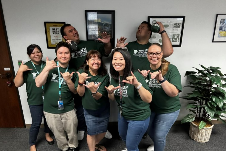 Alt text: A group of employees wearing matching green Hawaii Athletics shirts pose together in an office, making shaka gestures. Framed awards, sports memorabilia, and office plants are visible in the background, highlighting team spirit and community involvement.