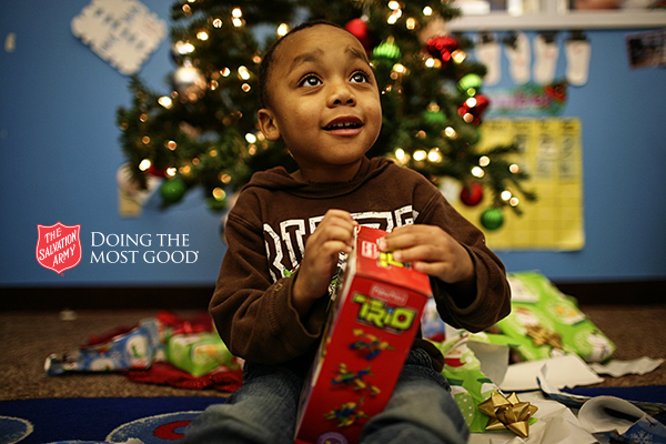 A child sits on the floor opening a holiday gift in front of a decorated Christmas tree, surrounded by wrapping paper and presents. The Salvation Army logo and the phrase “Doing the Most Good” appear on the left side of the image.