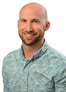 A professional headshot of a person with a shaved head wearing a light blue patterned aloha shirt, photographed against a light gray background.