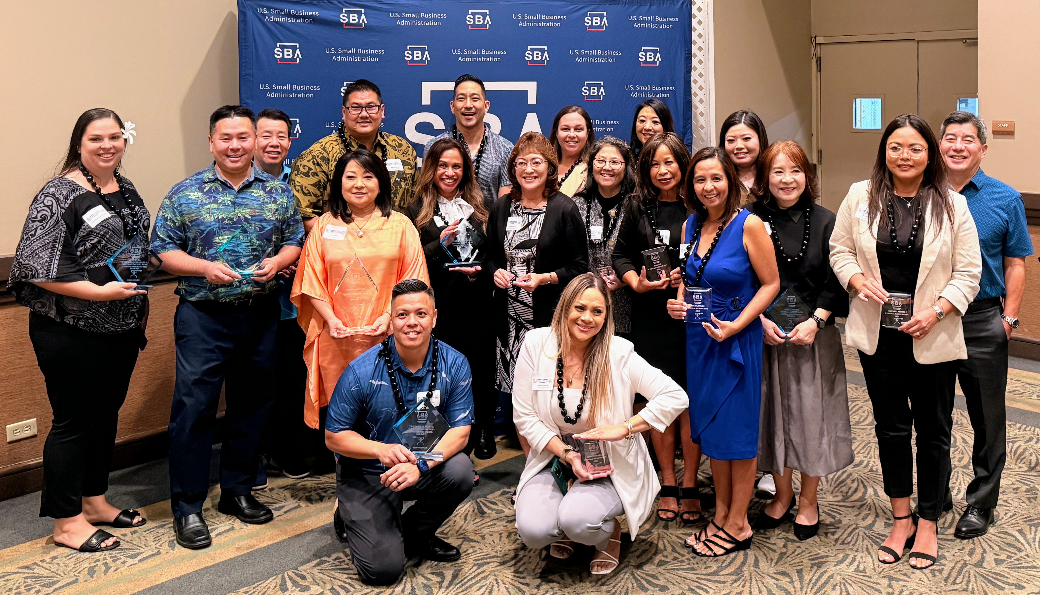 A large group of award recipients and business professionals pose for a photo in front of a U.S. Small Business Administration (SBA) backdrop. Several participants hold recognition plaques, highlighting an SBA awards and recognition event.