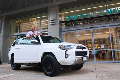 A person stands through the sunroof of a white SUV parked outside a Central Pacific Bank branch, holding a large ceremonial key. The bank entrance and downtown buildings are visible in the background, highlighting a vehicle giveaway or recognition event.