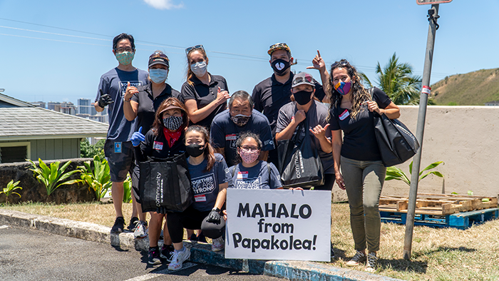 A group of volunteers wearing face masks pose outdoors behind a sign that reads “MAHALO from Papakōlea!” The group stands on a sunny residential street, many making shaka gestures while holding volunteer supplies and tote bags.