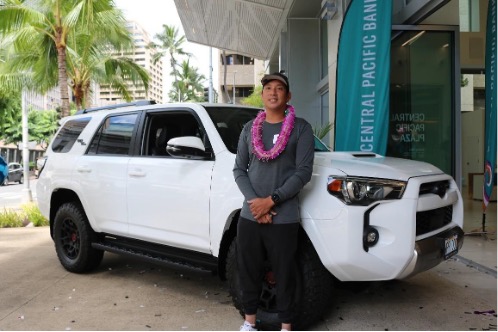 A person wearing a lei stands beside a white SUV outside a Central Pacific Bank branch. Palm trees, downtown buildings, and Central Pacific Bank signage are visible in the background.