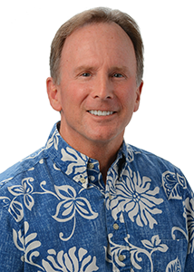 A professional headshot of a person wearing a blue aloha shirt with a white floral pattern, photographed against a light gray background.