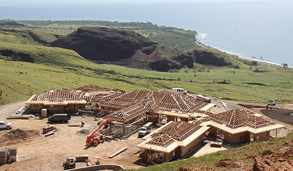 Aerial view of a large construction site with several buildings in various stages of framing, situated on a green coastal hillside. The Pacific Ocean, rugged cliffs, and surrounding landscape stretch into the background, highlighting the scenic location of the development.