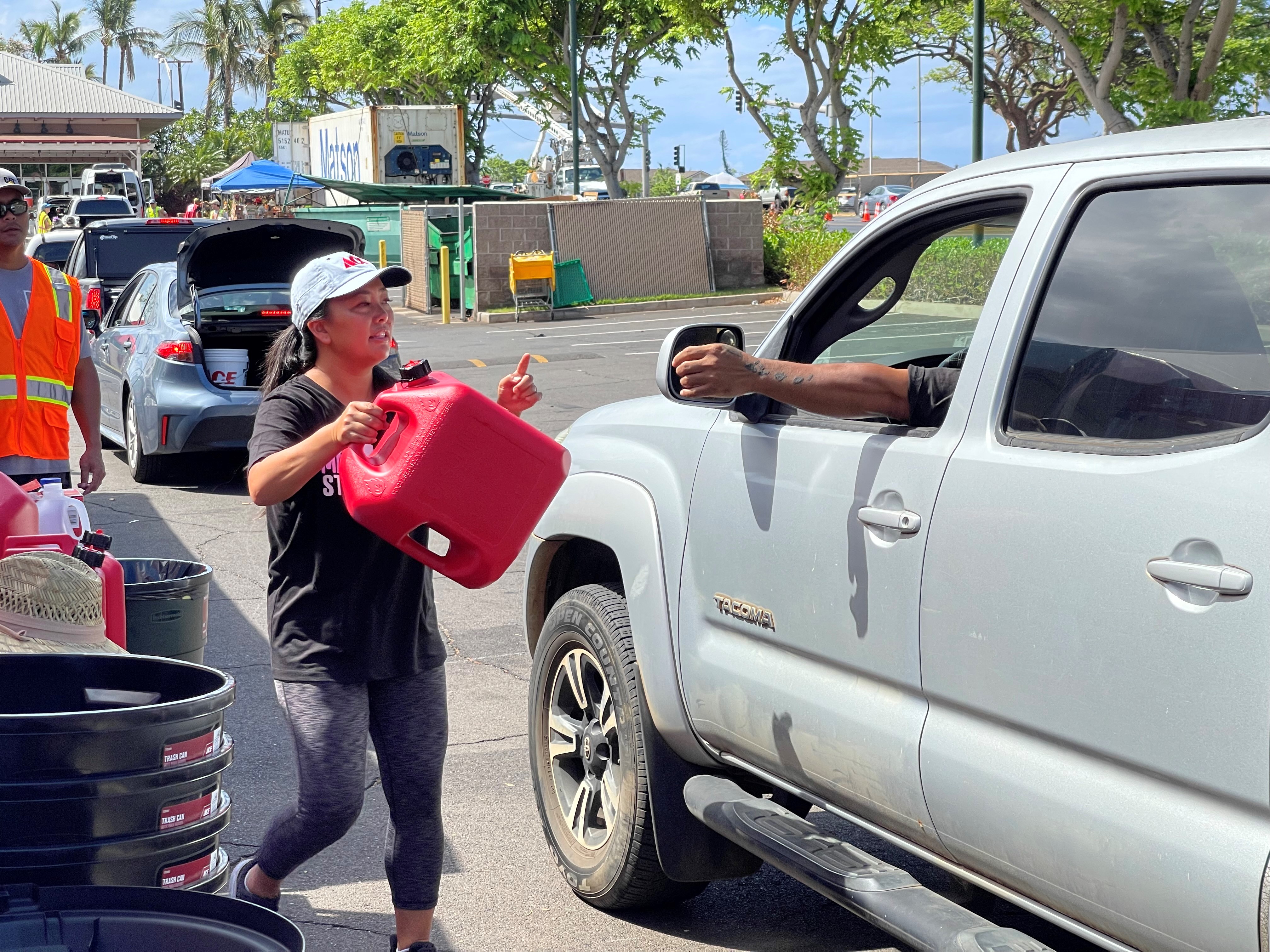 A volunteer carries a red fuel container to a pickup truck during a community distribution event in a parking lot. Other volunteers, vehicles, and supply stations are visible in the background as emergency preparedness items are handed out.
