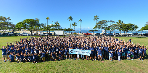 A large group of Central Pacific Bank employees gather on a grassy field for a group photo, holding a bank banner with palm trees, the ocean, and a clear blue sky in the background.