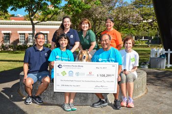 A group of seven people pose outdoors with a Collie dog and a large Central Pacific Bank donation check, seated and standing around a stone bench in a park setting.