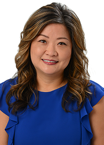 A professional headshot of a person wearing a royal blue blouse with ruffled sleeves, photographed against a light gray background.