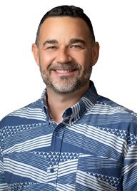 A professional headshot of a person wearing a red aloha shirt with a large white floral pattern, photographed against a light gray background.