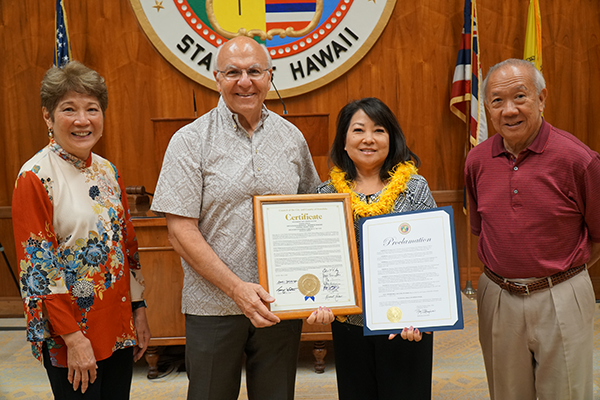 Four people stand in the Hawaiʻi State Capitol holding a certificate and proclamation during a recognition ceremony. The group poses in front of the State of Hawaiʻi seal, with U.S., Hawaiʻi, and other flags visible in the background.