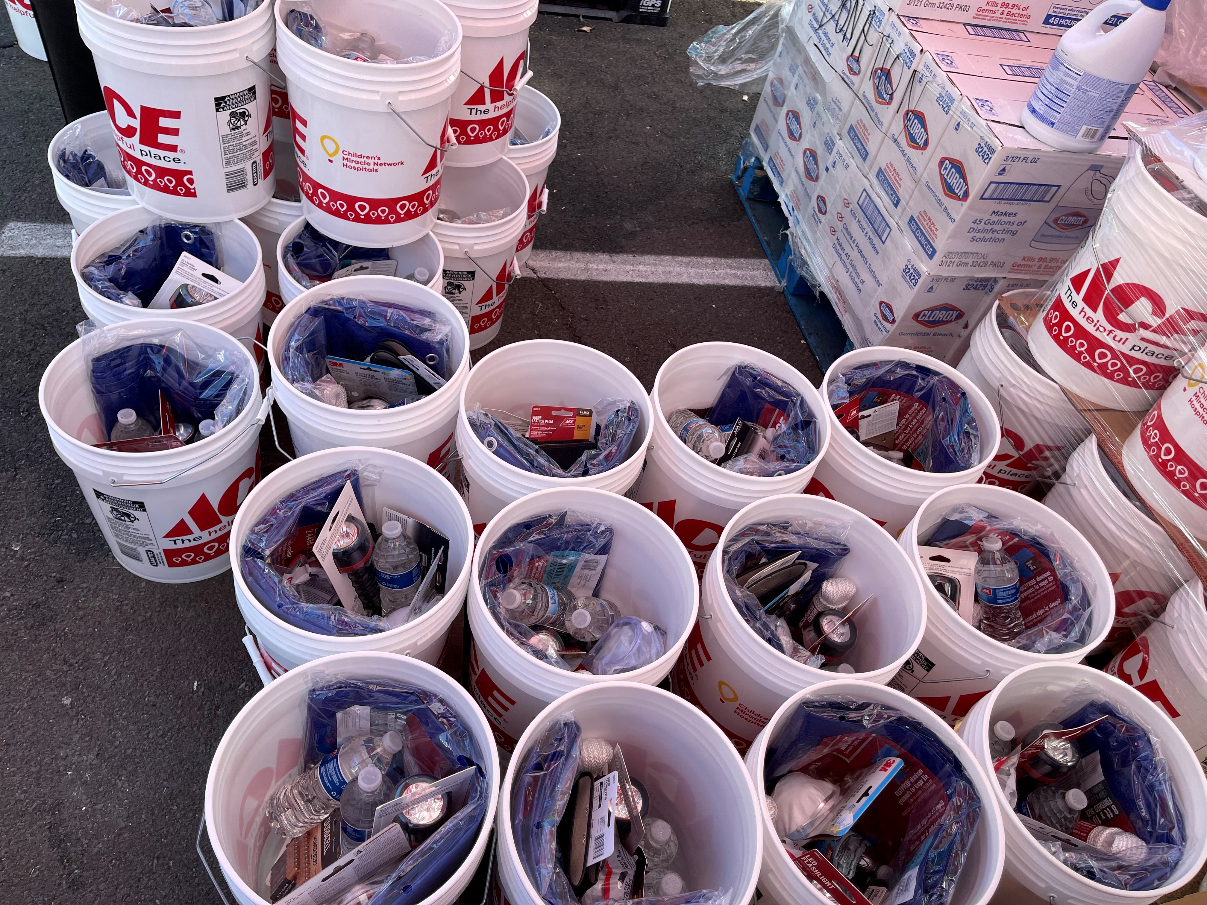 A collection of ACE-branded donation buckets filled with emergency and household supplies is arranged outdoors alongside boxes of cleaning products. The buckets contain items such as bottled water, flashlights, batteries, gloves, and other preparedness materials for community relief or disaster response efforts.