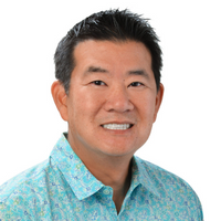 A professional headshot of a person wearing a light blue aloha shirt with a subtle floral pattern, photographed against a light gray background.
