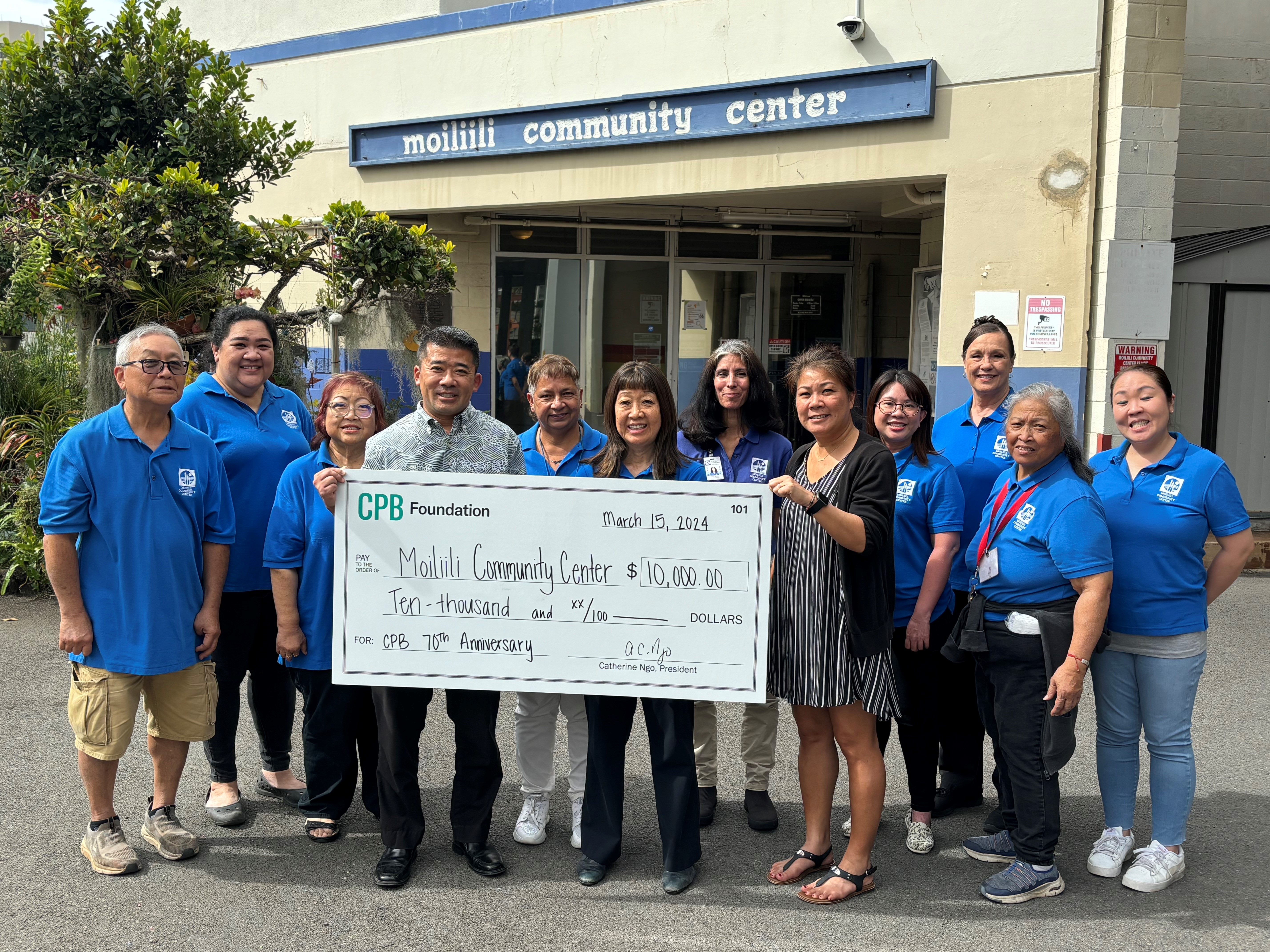 A group of community members stands outside the Moiliili Community Center holding a $10,000 CPB Foundation donation check. The participants pose in front of the center’s entrance during a community support and grant presentation event.