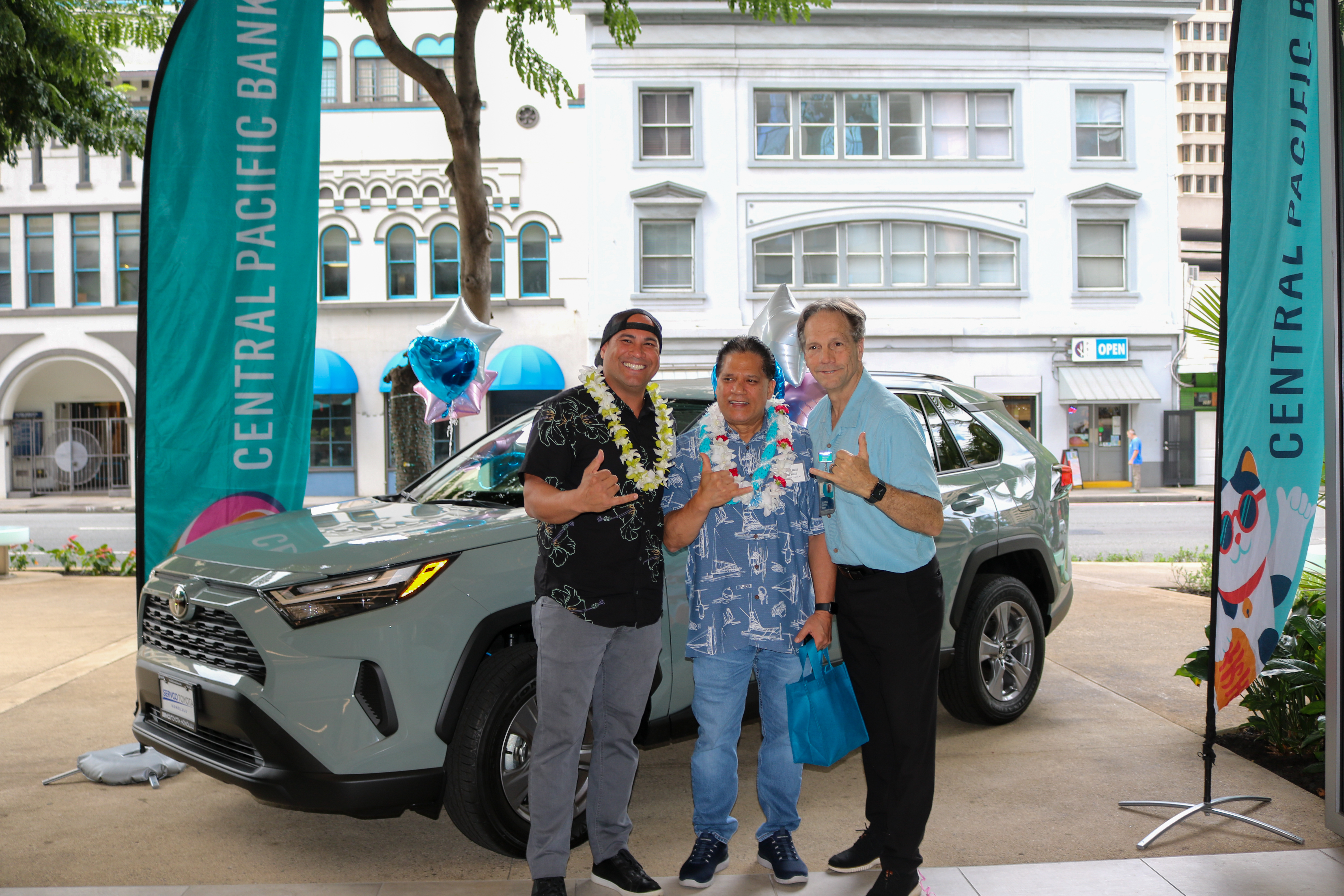Three people wearing flower leis pose in front of a Toyota RAV4 outside a Central Pacific Bank branch. Central Pacific Bank banners and decorative balloons frame the vehicle, highlighting a promotional car giveaway event.