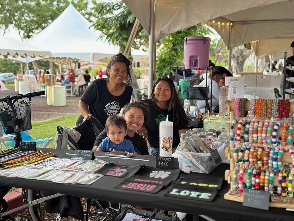 A group of adults and a young child sit and stand behind a vendor table displaying colorful handmade bracelets, pouches, lanyards, and other crafts at an outdoor market. The booth is set up under a tent, with other vendors and visitors visible in the background.