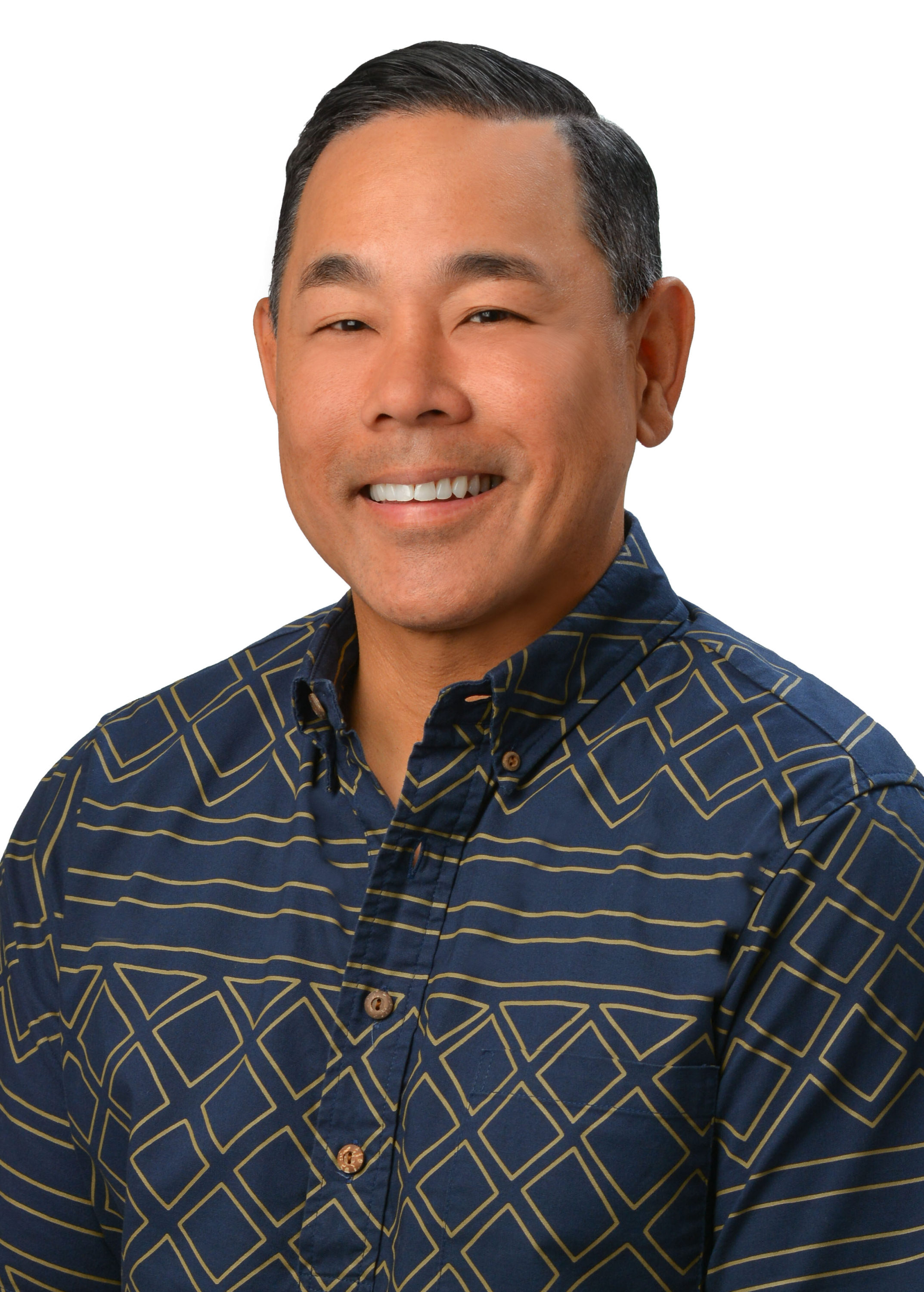 A professional headshot of a person wearing a blue aloha shirt with a geometric gold pattern, photographed against a light gray background.
